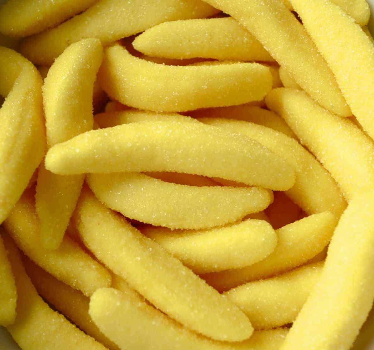 Fizzy banana sweets in a bright yellow, shaped like small banana figures, with a tangy, fizzy coating, scattered on a white surface.